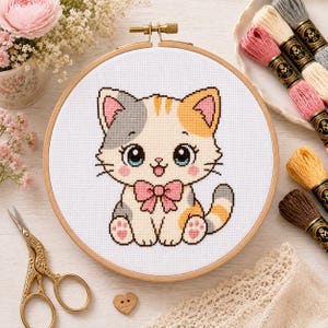 May include: A cross-stitch embroidery project in a round wooden hoop featuring a cartoon kitten with white, gray, and orange fur, large blue eyes, and a pink bow tie. The design is stitched on white Aida fabric with a grid pattern visible.