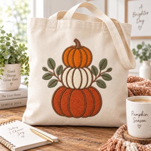 May include: A cream-coloured canvas tote bag featuring an embroidered design of three stacked pumpkins in orange, white, and rust colours, accented with green leaves on either side. The bag has two sturdy fabric handles and a simple, clean aesthetic.