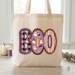 Könnte beinhalten: Eine beige Tragetasche aus Segeltuch mit einer Stickerei des Wortes BOO. Der Buchstabe B hat ein schwarz-weißes Karomuster, das O ist lila mit weißen Geistern und Fledermäusen, und das zweite O hat vertikale orange, lila und weiße Streifen.