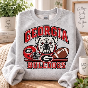 May include: A light gray crewneck sweatshirt featuring a graphic print with the text GEORGIA BULLDOGS in red and black. The design includes a white bulldog face, a red football helmet with a black G logo, and a brown football with white laces.