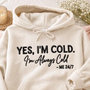 Puede incluir: Una sudadera con capucha de color crema que presenta un texto bordado en negro que dice: YES, I'M COLD. I'm Always Cold - ME 24/7. La prenda incluye un cordón a juego, un bolsillo tipo canguro y mangas largas con puños acanalados.