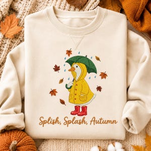 May include: A cream-colored crewneck sweatshirt featuring an embroidered design of a white goose wearing a yellow raincoat and red rain boots while holding a green umbrella. Falling autumn leaves and blue raindrops surround the goose, with the text Splish, Splash, Autumn printed in brown below the graphic.