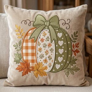 Puede incluir: Un cojín cuadrado de color beige con un diseño de calabaza bordado. La calabaza está dividida en tres secciones con cuadros vichy naranjas, un estampado floral y una sección verde con corazones, rematada con un lazo verde y hojas de otoño.