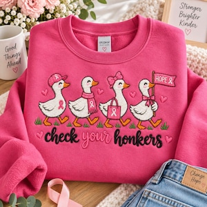 Puede incluir: Una sudadera de cuello redondo rosa brillante con un diseño bordado de cuatro patos blancos. Los patos llevan accesorios rosas y sostienen una bandera con el texto "HOPE". Debajo de los patos, el texto "check your honkers" está bordado en negro.