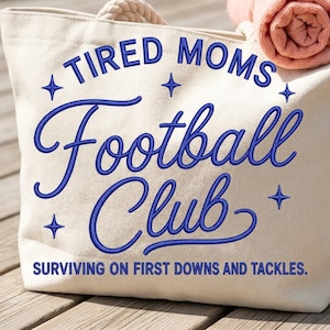May include: A cream-colored canvas tote bag featuring blue embroidered text that reads TIRED MOMS Football Club and SURVIVING ON FIRST DOWNS AND TACKLES. The design includes small blue star accents and thick rope handles at the top of the bag.