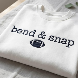 Op de afbeelding: Een wit sweatshirt met ronde hals en donkerblauw geborduurde tekst bend & snap. Onder de tekst bevindt zich een klein, donkerblauw geborduurd American football-icoon met witte stikseldetails, gecentreerd op het borstgedeelte van het kledingstuk.