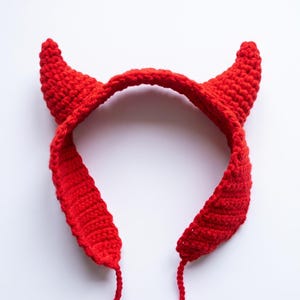 Può includere: Una fascia all'uncinetto rosso brillante caratterizzata da due protuberanze appuntite a forma di corno rivolte verso l'alto. L'accessorio è realizzato con un motivo a maglia visibile e include sottili lacci all'uncinetto alle estremità.