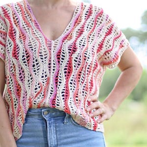 May include: A short-sleeved crochet top featuring a V-neckline and a vertical wavy pattern in shades of cream, pink, and orange, paired with light blue denim jeans. The garment has an open-knit texture with repeating vertical columns.