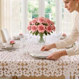 May include: A white rectangular tablecloth featuring an intricate crochet lace pattern with floral motifs. The tablecloth is draped over a dining table set with white plates, silverware, clear glassware, and a glass vase filled with pink roses.
