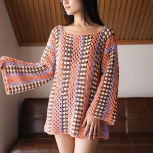 May include: A long-sleeved crochet sweater featuring a vertical striped pattern in shades of orange, lavender, and dark brown. The garment has a wide scoop neckline and flared sleeves, constructed with a repeating open-work granny square stitch.