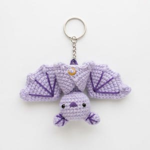 May include: A light purple crocheted bat keyring featuring dark purple embroidered wing details, a small dark purple nose, black bead eyes, and a circular gold-toned charm with a crescent moon and star design attached to the top of the body.