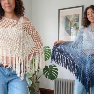 Puede incluir: Dos ponchos de ganchillo con un patrón de punto de red abierto y largos flecos en los bordes inferiores. Un poncho es de color crema sólido, mientras que el otro muestra un degradado azul que va del azul claro en la parte superior al azul marino.