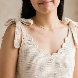 May include: A cream-colored crochet tank top featuring a scalloped neckline and shoulder straps that tie into bows. The garment is crafted with a fine, uniform stitch pattern, providing a textured appearance across the front and shoulder areas.