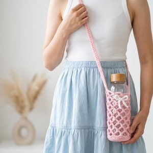 May include: A light pink crocheted water bottle holder with a long shoulder strap and a small white bow detail. The holder features an open lattice pattern and contains a clear glass water bottle with a light brown wooden cap.