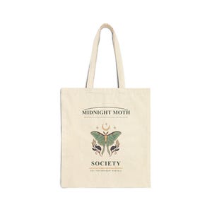 Könnte beinhalten: Eine cremefarbene Stofftasche aus Segeltuch mit einer zentralen Grafik eines grünen Nachtfalters und einer Mondsichel darüber. Der Text MIDNIGHT MOTH SOCIETY und EST. FOR ORDINARY MARVELS ist in schwarzer Schrift auf der Vorderseite zu sehen.