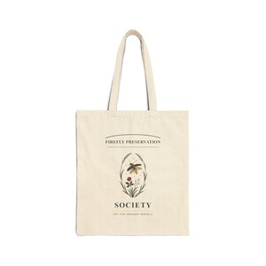 Könnte beinhalten: Eine beigefarbene Tragetasche aus Segeltuch mit einer zentralen Grafik eines Glühwürmchens, umgeben von botanischen Elementen in einem ovalen Rahmen. Der Text FIREFLY PRESERVATION SOCIETY und EST. FOR ORDINARY MARVELS steht in Schwarz.