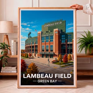 May include: A framed art print of Lambeau Field in Green Bay, featuring the stadium exterior with brick and green accents, autumn trees, and a blue sky. The text reads LAMBEAU FIELD and GREEN BAY, with stadium signage for LAMBEAU FIELD.