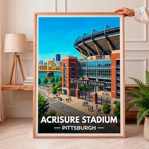 Op de afbeelding: Een ingelijste kunstdruk met een levendige illustratie van het Acrisure Stadium in Pittsburgh. De afbeelding toont de stadionstructuur, een gele brug over een rivier en stadsgebouwen onder een helderblauwe lucht. De tekst luidt Acrisure Stadium Pittsburgh.