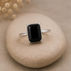 May include: A silver-toned ring featuring a large, rectangular, faceted black gemstone set in a four-prong setting. The band is smooth and polished, connecting to the central stone setting, which holds the dark, opaque rectangular gem securely.