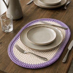 Puede incluir: Un mantel individual de ganchillo de forma ovalada con un centro de color crema y un borde grueso de color lavanda. El mantel está dispuesto con un plato llano beige grande, un plato de ensalada beige más pequeño, un tenedor y un cuchillo de acero inoxidable.