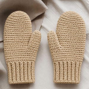 Op de afbeelding: Een paar beige gehaakte wanten met een dicht, gestructureerd steekpatroon over het hoofdgedeelte en een geribbelde boord bij de pols. De wanten zijn ontworpen met een afgeronde bovenkant en een apart duimgedeelte aan de binnenzijde.