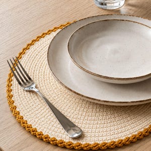 May include: A round, cream-colored crocheted placemat with a mustard yellow scalloped border. The placemat holds a speckled, off-white ceramic dinner plate and a smaller matching bowl, topped with a polished stainless steel dinner fork.