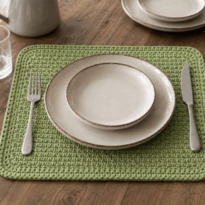 May include: A rectangular light green crocheted placemat featuring a textured stitch pattern. Centered on the placemat are two stacked off-white ceramic plates with brown rims. A silver fork is placed to the left and a silver knife to the right.