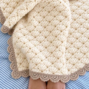 May include: A cream-coloured crochet blanket featuring a repeating shell stitch pattern throughout the main body. The blanket is finished with a decorative, scalloped border in a contrasting light brown yarn, creating a textured edge.