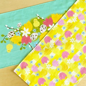 May include: Two fabric placemats with a yellow and pink checkered pattern and a green placemat with a floral design and yellow chicks. The yellow and pink placemat has a repeating pattern of pink eggs and yellow flowers.