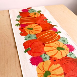 May include: A white table runner with a autumn-themed design featuring a row of orange, yellow, and green pumpkins with pink and green floral accents.