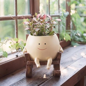 以下が含まれることがあります： シンプルな赤い笑顔と2本の小さな腕を持つ、丸いキャラクターの形をしたクリーム色の陶器製プランター。茶色の木製ミニチュアロッキングチェアの上に置かれています。プランターには、緑とピンクの斑入りの葉を持つ小さな植物が植えられています。