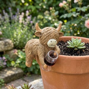 May include: A tan Highland cow figurine with textured fur, horns, and a cream-colored muzzle, designed to hang on the rim of a terracotta pot. The cow is holding a small, clear glass filled with dark red liquid, positioned next to a green succulent.