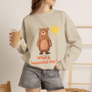Puede incluir: Una sudadera beige de manga larga con una ilustración de un oso marrón, un gráfico de un sol amarillo y el texto naranja "What a beautiful day!" impreso en el frente. La sudadera tiene cuello redondo y mangas largas con puños acanalados.