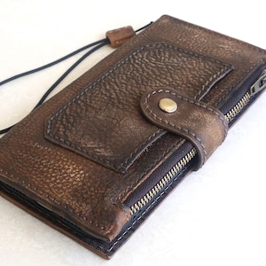 May include: A brown textured leather wallet featuring a rectangular front pocket, a metal snap button closure, and a side zipper compartment. A thin black elastic wrist strap is attached to the top corner of the wallet, which has visible stitching.