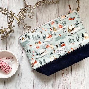 May include: A quilted cosmetic bag featuring a winter village scene in light blue, red, and white. The base is a dark blue fabric. A small white bowl with red and white twine and a decorative sprig are also in the image.
