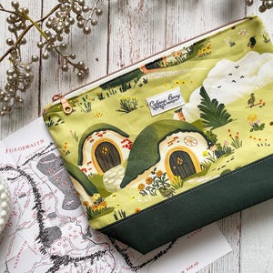 May include: A rectangular, green and yellow fabric pouch with a landscape design featuring hobbit-hole houses. The pouch has a dark green bottom, a gold zipper, and a white label with text. A map and a small white bowl are next to the pouch.