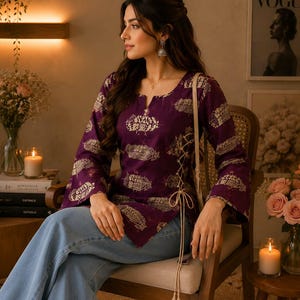 Op de afbeelding: Een donkerpaarse tuniek met lange mouwen, voorzien van een herhalend goudkleurig paisley-patroon, een split-halslijn en een zijdelingse vetersluiting met beige koorden. Het kledingstuk wordt gecombineerd met een lichtblauwe spijkerbroek.
