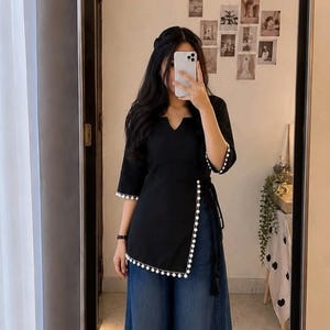 May include: A black wrap-style tunic featuring a V-neckline, three-quarter length sleeves, and white pom-pom trim along the edges. The tunic is paired with wide-leg blue denim jeans and white sneakers, creating a casual and layered ensemble.