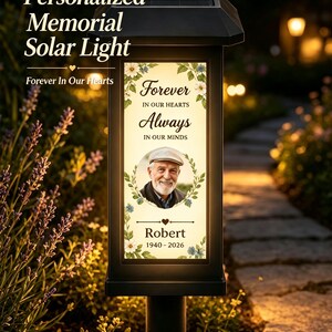 May include: A black solar-powered memorial light featuring a rectangular illuminated panel with floral accents. The text reads Forever IN OUR HEARTS Always IN OUR MINDS, with a circular portrait of a man, the name Robert, and the years 1940-2026.