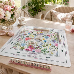 Pode incluir: Um tapete de Mahjong quadrado com fundo branco e um padrão floral colorido de flores rosas, azuis e amarelas. O tapete inclui instruções de jogo impressas e texto como Charleston, Dragons e Jokers. Um suporte contém peças de Mahjong.