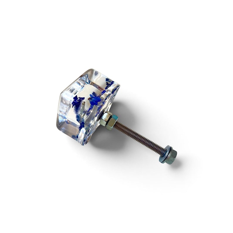 Op de afbeelding: Een vierkante meubelknop van heldere hars met ingebedde gedroogde blauwe bloemen. De knop is voorzien van een lange metalen bout met schroefdraad aan de achterkant, vastgezet met twee zilverkleurige metalen moeren en een ring voor installatie.