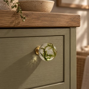 May include: A spherical clear resin cabinet knob featuring embedded green moss, attached to a gold-toned metal base. The knob is mounted on a sage green wooden drawer front with a visible wood grain texture and a horizontal panel design.