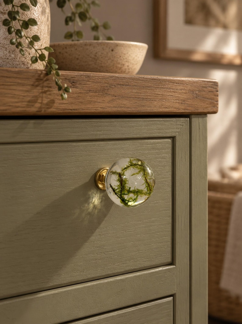 May include: A spherical clear resin cabinet knob featuring embedded green moss, attached to a gold-toned metal base. The knob is mounted on a sage green wooden drawer front with a visible wood grain texture and a horizontal panel design.