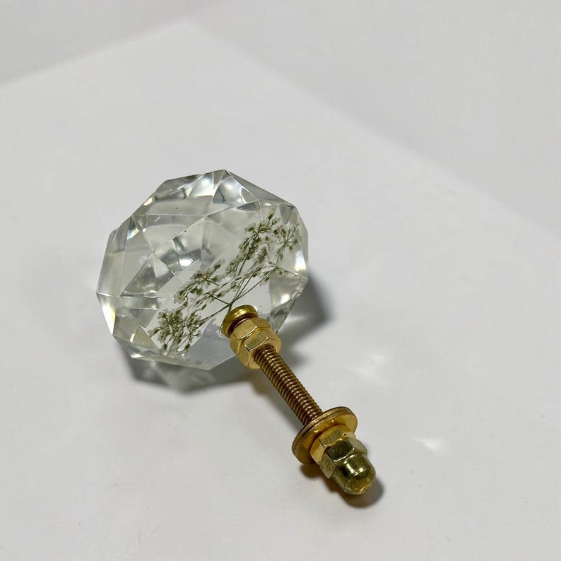 May include: A clear, faceted, diamond-shaped cabinet knob made of resin, containing a delicate, dried white flower inside. The knob is attached to a gold-toned metal bolt with a matching washer and nut for secure installation on furniture.