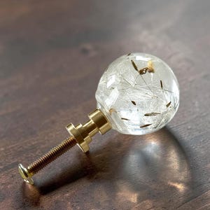 May include: A spherical cabinet knob featuring a clear resin orb containing delicate dandelion seeds. The knob is attached to a metallic gold-toned base and a threaded screw, providing a functional hardware piece for furniture or cabinetry.