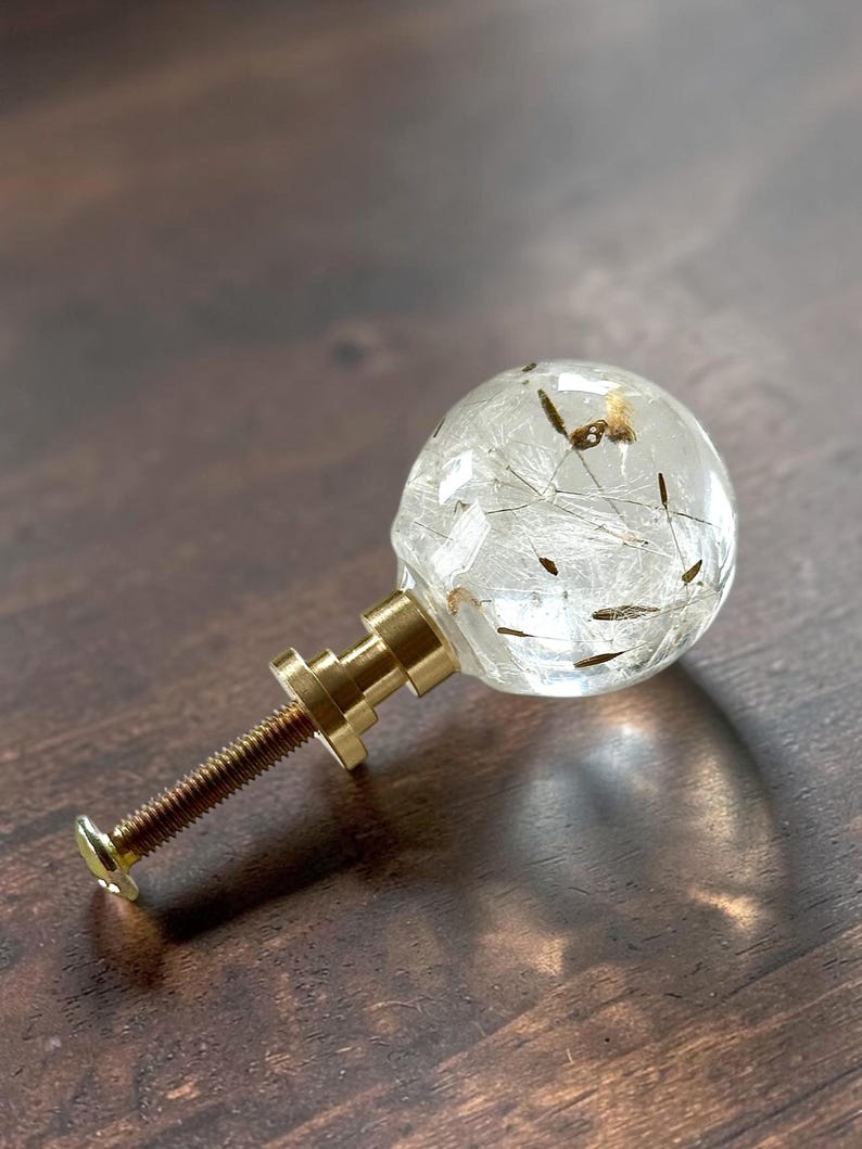 May include: A spherical cabinet knob featuring a clear resin orb containing delicate dandelion seeds. The knob is attached to a metallic gold-toned base and a threaded screw, providing a functional hardware piece for furniture or cabinetry.