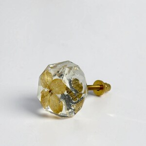 May include: A clear, faceted, hexagonal resin cabinet knob featuring an embedded dried yellow flower and small gold-toned metallic flakes. The knob is attached to a brass-colored metal bolt with a matching nut extending from the back side.