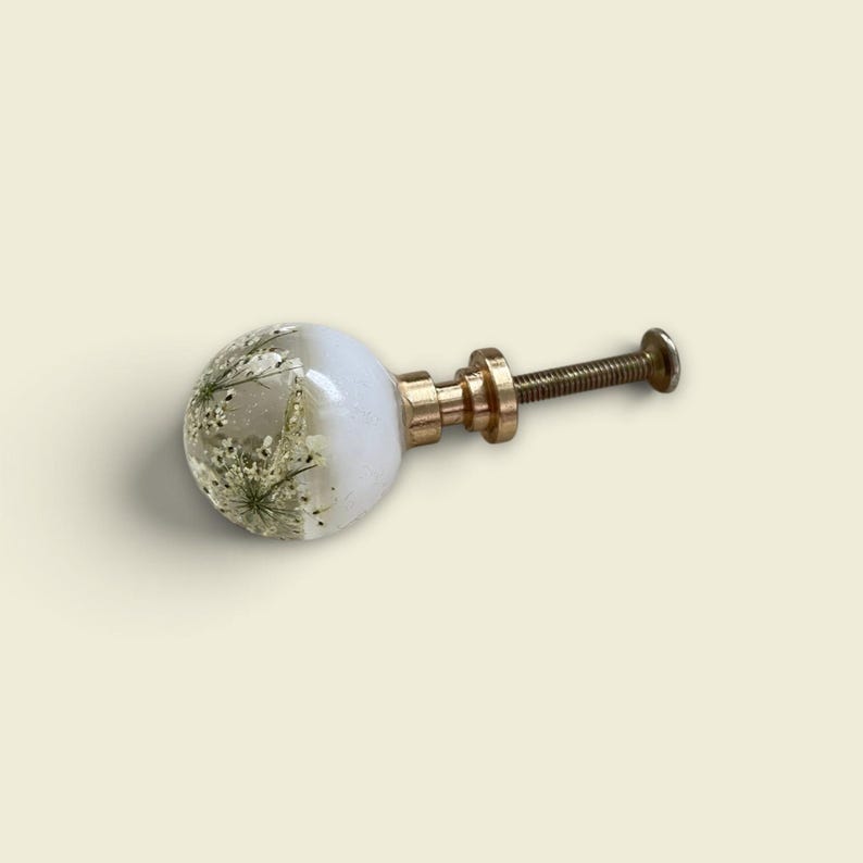 May include: A spherical cabinet knob featuring a clear resin dome embedded with dried white Queen Anne's lace flowers. The knob has a smooth, opaque white base and is attached to a gold-toned metal post with a threaded screw and a flat circular end.
