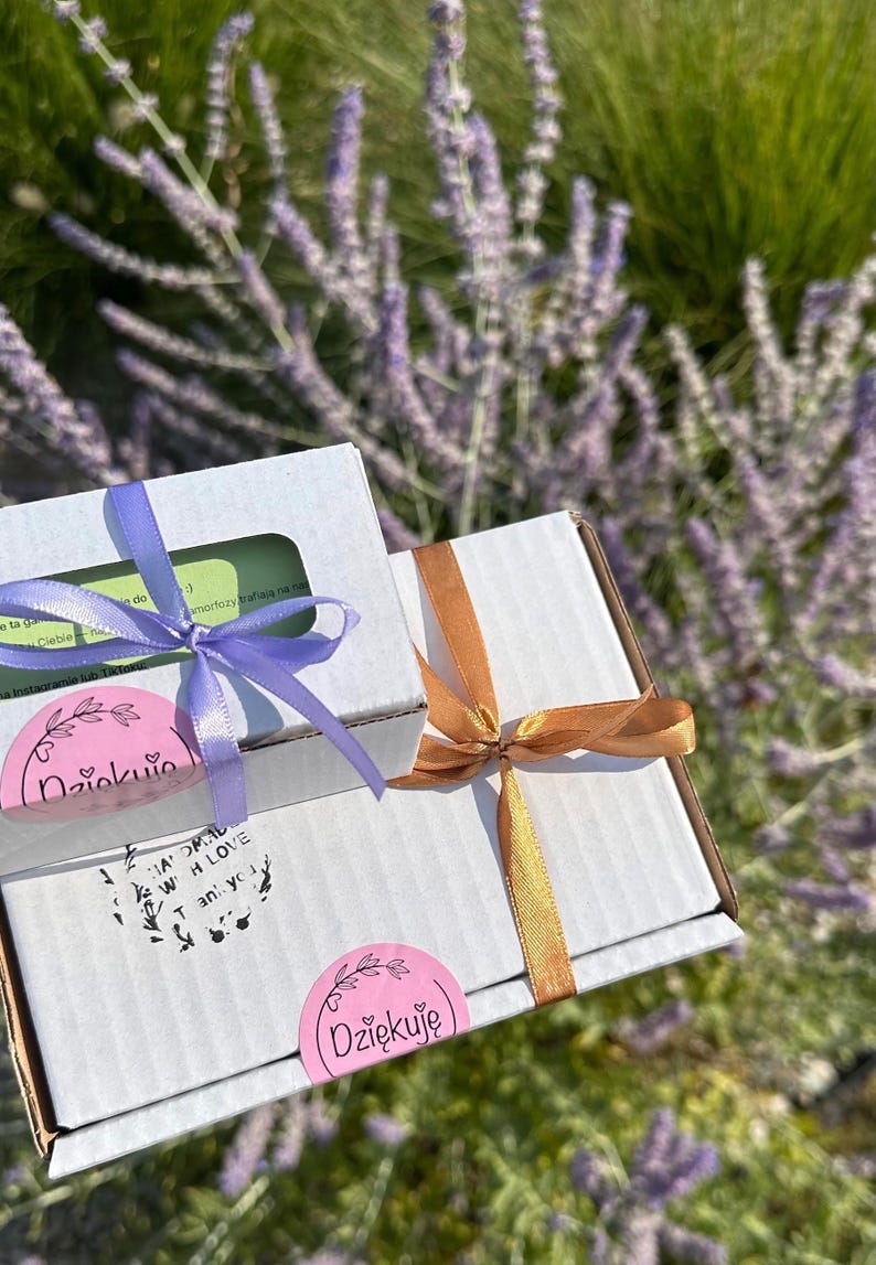 May include: Two white cardboard shipping boxes tied with ribbon. The smaller box has a light purple ribbon and a pink sticker reading Dziękuję. The larger box has a bronze ribbon and a pink sticker reading Dziękuję, plus text reading Handmade with love Thank you.
