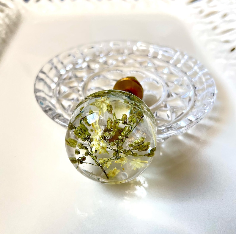 K&ouml;nnte beinhalten: Ein kugelf&ouml;rmiger Knauf aus klarem Harz mit eingebetteten getrockneten gelben und gr&uuml;nen Blumen. Der Knauf hat einen goldfarbenen Metallsockel und eine glatte, polierte Oberfl&auml;che, die die zarten botanischen Details im Inneren sichtbar macht.